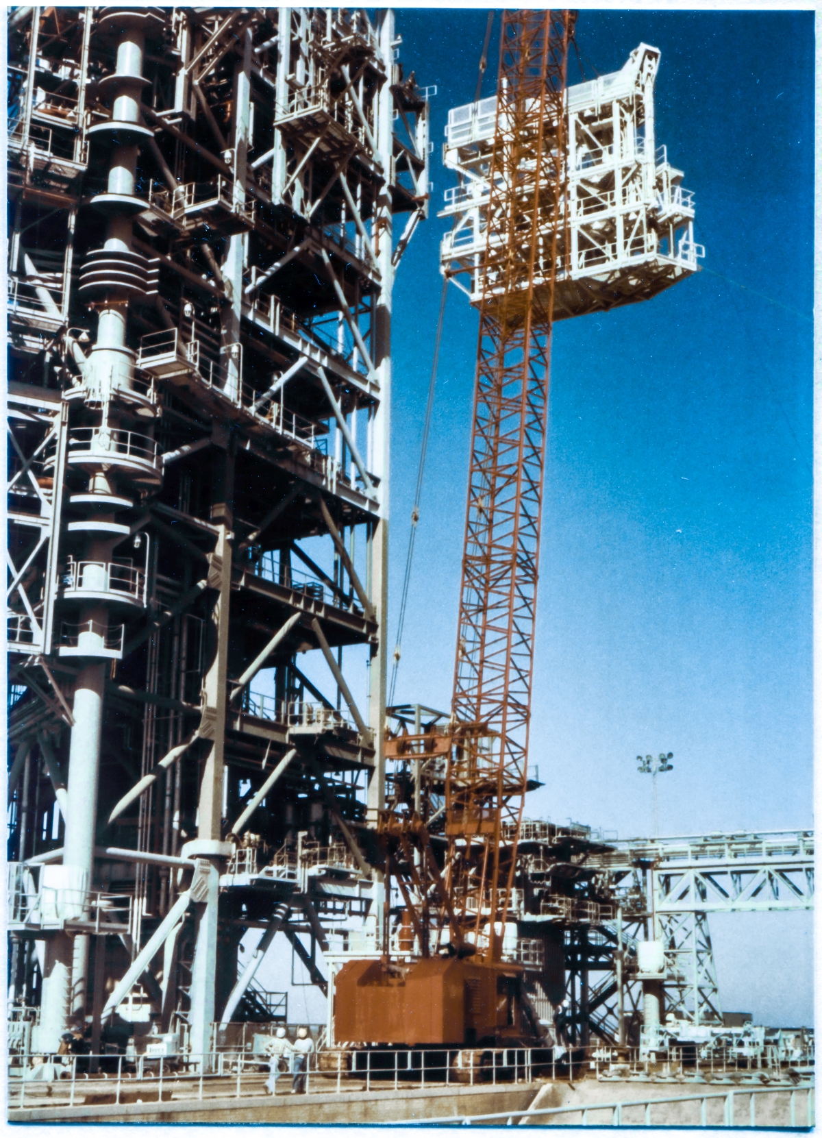 Image 123. At Space Shuttle Launch Complex 39-B, Kennedy Space Center, the IAA (Intertank Access Arm) has risen roughly a hundred and fifty feet above the Pad Deck, and is nearing its final location, bolted to its Support Structure, which remains just above it fifteen feet or so, immediately to its left. The Support Structure extends a bit out and to the right, beyond the north side of the FSS, which dominates the whole left side of the frame. Standing on top of the Support Structure, at the 220'-0” level, a Union Ironworker from Local 808, working for Ivey Steel, can be seen at the edge of the precipice, ready to begin the process of connecting the IAA to the tower with A325 high-strength structural bolts when it arrives. Left of him, partially blocked by the large and complex connection weldment attached to the FSS Side 1-4 Corner Column, the silhouette of another ironworker, in shadow, also at the edge of the precipice, can be seen. Photo by James MacLaren.